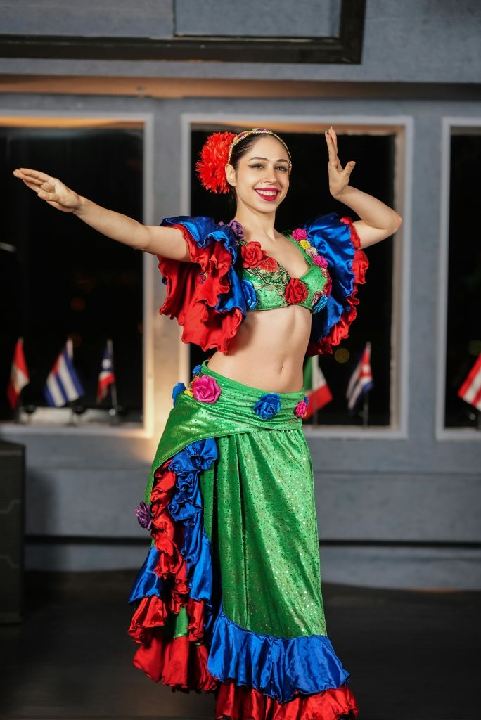 두 팔을 쭉 뻗은 채 화려하고 주름진 의상 포즈를 취한 미소를 짓는 여성. 배경에 깃발이 보입니다.