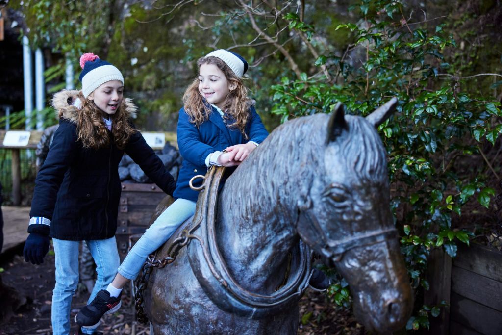 겨울 옷을 입은 두 어린이가 야외에서 말의 대형 조각상과 상호 작용합니다.