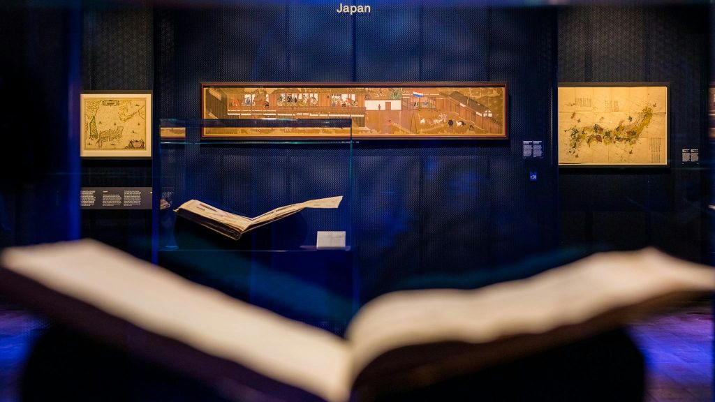 짙은 파란색 벽에 일본 두루마리 그림을 전시한 박물관 전시물과 두 권의 열린 책이 전면에 전시되어 있습니다.