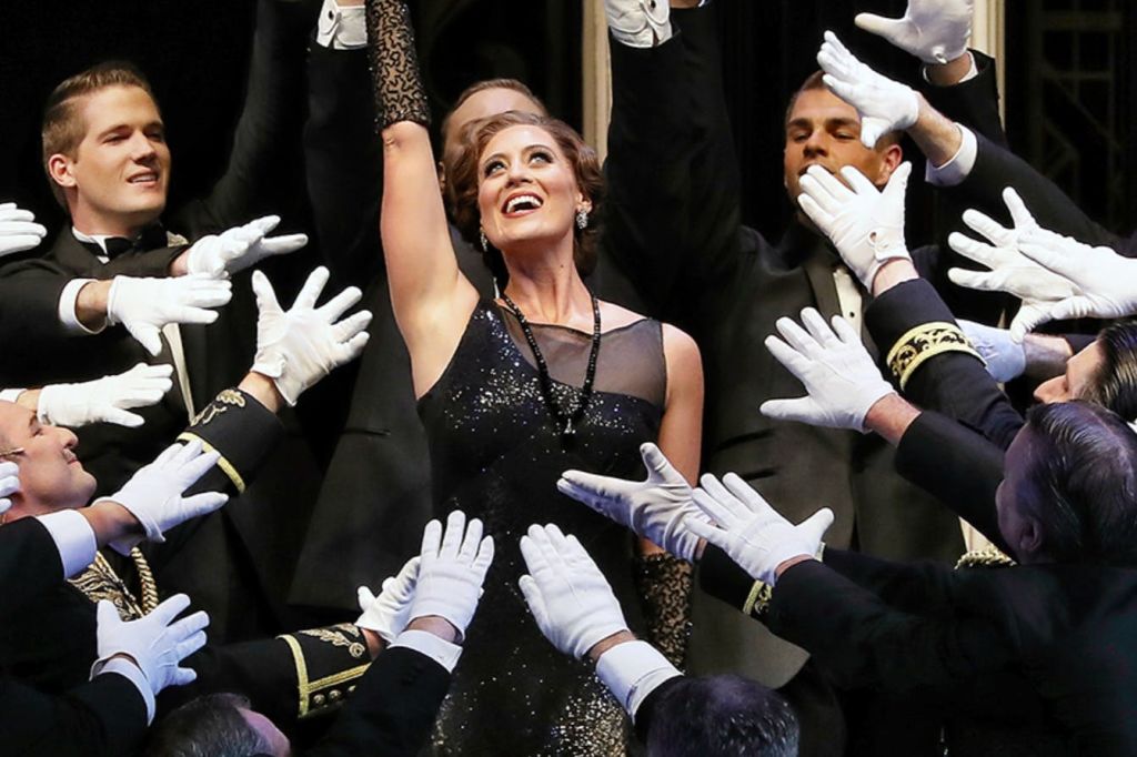 반짝이는 검은색 드레스를 입은 한 여성이 자신을 향해 장갑을 낀 손을 뻗은 정장 차림의 사람들에게 둘러싸인 채 팔을 들고 있습니다.