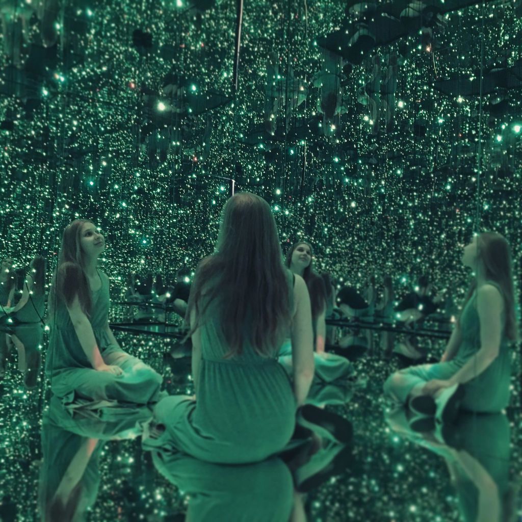 녹색 드레스를 입은 여성이 녹색 불빛이 가득한 거울 방에 다리를 꼬고 앉아 여러 개의 반사를 연출합니다.