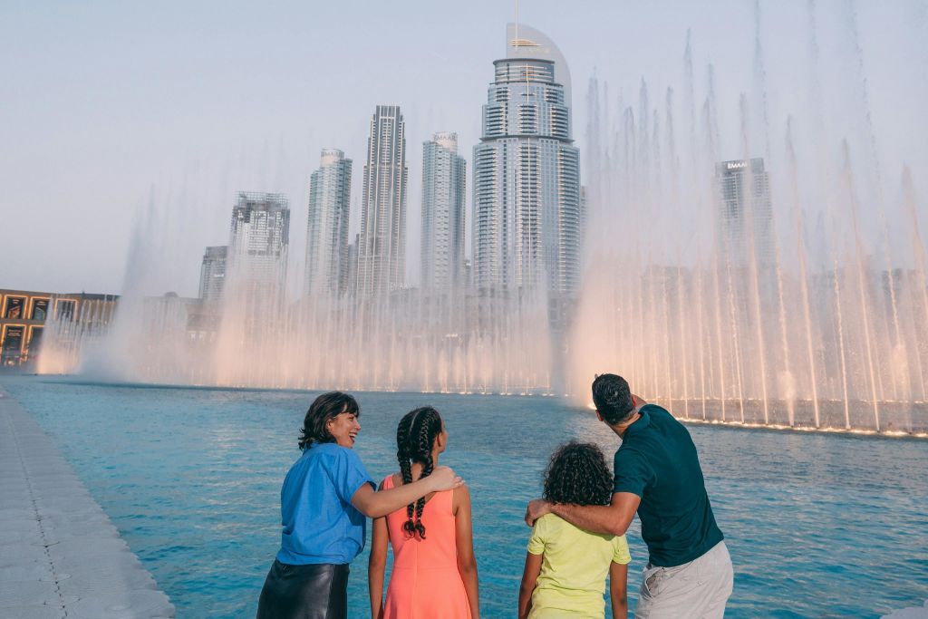 4인 가족이 높은 도시 건물을 배경으로 수영장 옆의 분수 디스플레이를 감상하고 있습니다.