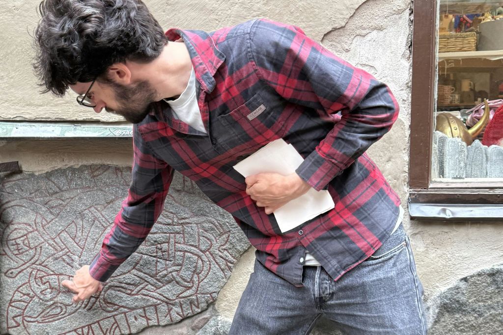 빨간색 격자무늬 셔츠를 입은 남성이 정교한 문양이 새겨진 돌을 가리키며 흰 종이를 들고 있습니다.