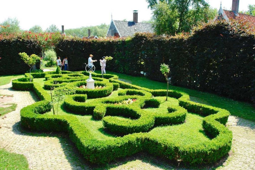 잘 꾸며진 울타리 미로, 흰색 동상, 탐험하는 여러 사람, 건물을 배경으로 한 정원.