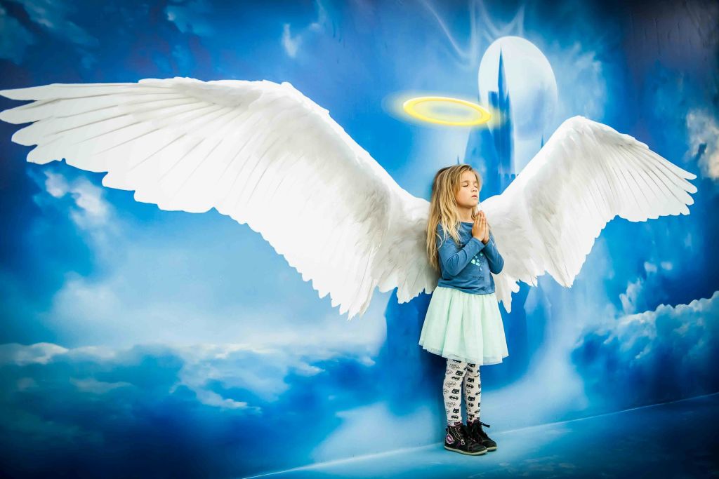 커다란 흰색 천사 날개와 후광을 가진 소녀가 하늘을 테마로 한 배경에 서서 손을 잡고 기도하고 있습니다.