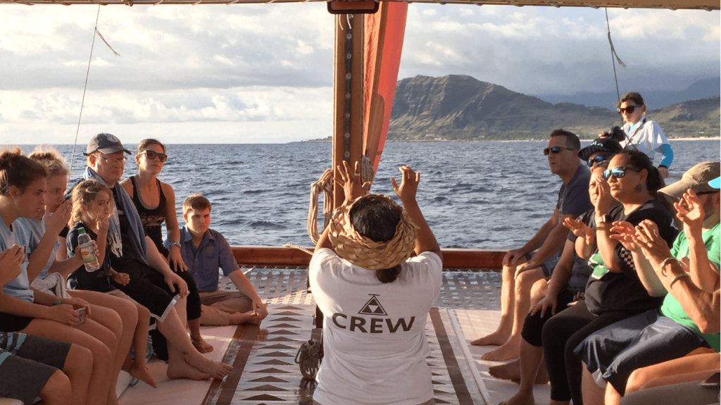 한 무리의 사람들이 보트에 앉아 박수를 치고 있는데, 앞에 승무원이 있고 저 멀리 산이 있습니다.