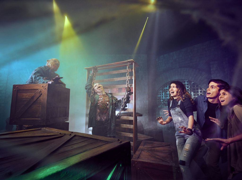 세 사람은 어둡고 안개가 자욱한 방 안에서 극적인 조명 아래 사슬로 묶인 해골 소품으로 두려움과 흥분으로 반응합니다.