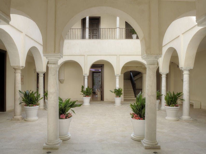 아치형 기둥, 화분, 계단, 문이 있는 위쪽 발코니가 있는 안뜰입니다.