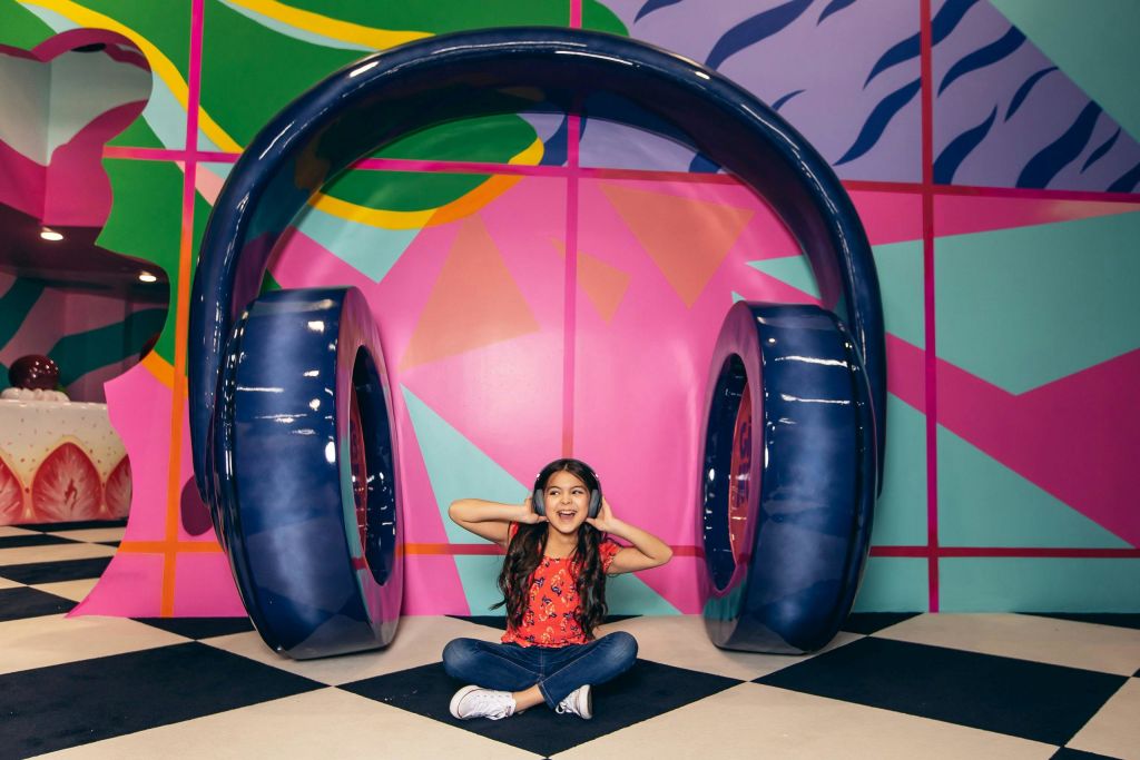 한 소녀가 커다란 파란색 헤드폰 조형물 아래 다리가 교차된 채 뺨에 손을 얹고 화려한 무늬를 배경으로 미소 짓고 있습니다.