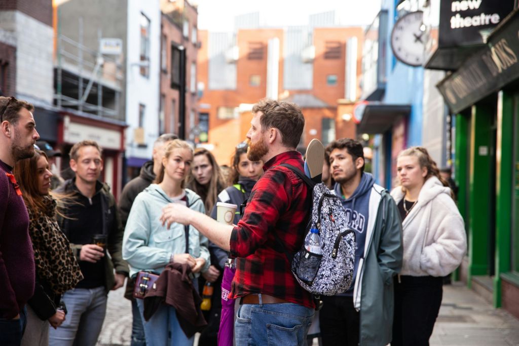빨간색 격자무늬 셔츠를 입은 남성이 도시 거리에 있는 한 무리의 사람들과 컵을 들고 스케이트보드가 달린 배낭을 메고 이야기하고 있습니다.