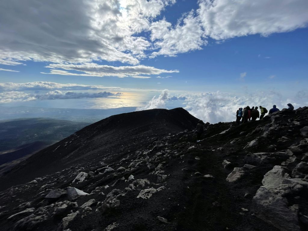 먼 바다가 내려다보이는 부분적으로 흐린 하늘 아래 바위가 많은 산비탈에 여러 사람이 서 있습니다.