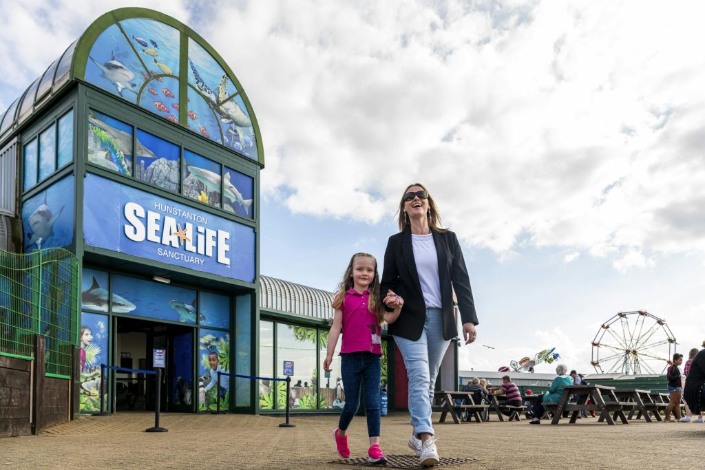 한 여성과 어린이가 Hunstanton SEA LIFE Sanctuary 밖에서 손을 잡고 걷고 있습니다. 다른 방문객과 관람차가 보입니다.
