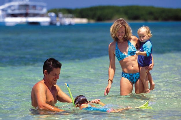 두 자녀를 둔 남녀가 해변에서 얕고 맑은 물을 즐깁니다. 한 아이는 스노클링을 하고 다른 아이는 여성이 잡습니다.