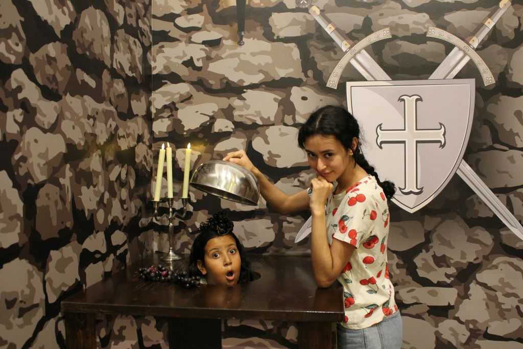 체리 무늬 셔츠를 입은 여성이 테이블 위로 은색 돔을 들고 있으며, 어린아이의 머리가 표면을 뚫고 나타나 놀란 표정을 짓고 있습니다. 배경에는 위장 디자인과 검 엠블럼이 있는 방패가 있습니다.