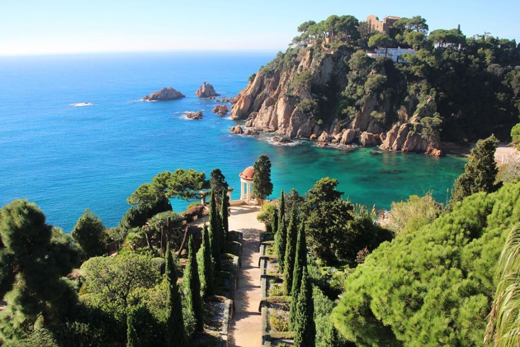 절벽, 청록색 물, 작은 정자로 이어지는 편백나무가 늘어선 정원길이 있는 지중해 해안 풍경.