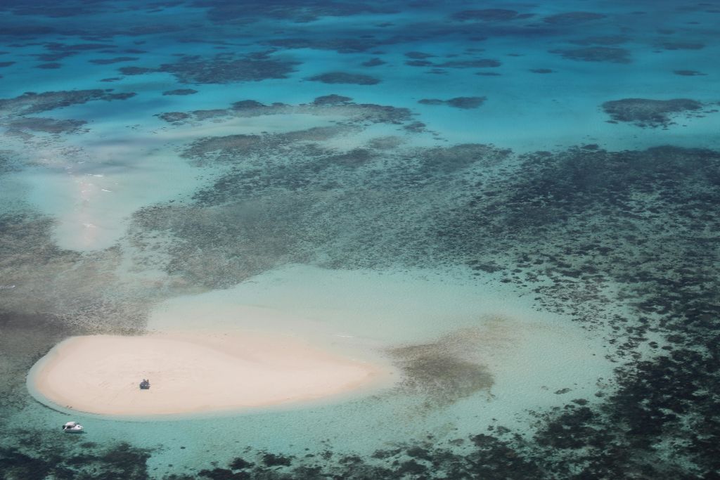 산호초가 보이는 청록색의 얕은 바다로 둘러싸인 외딴 모래 섬의 공중 전망.
