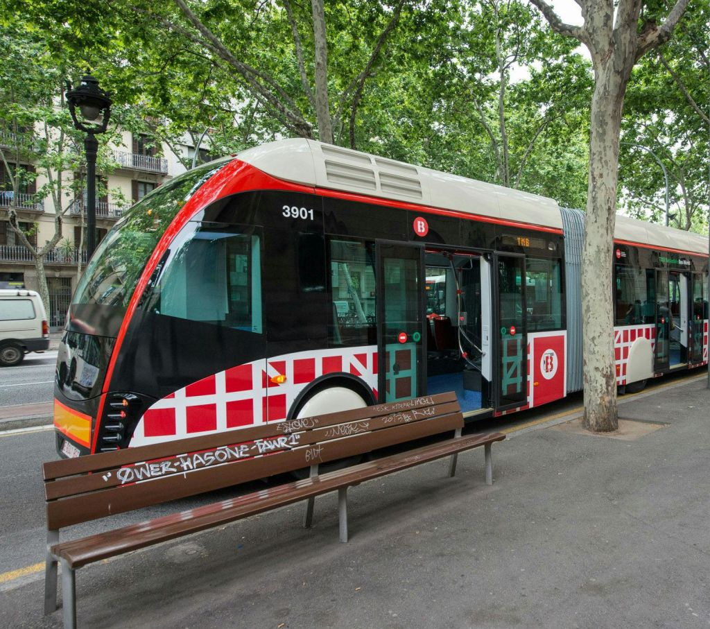 빨간색과 흰색으로 마디가 있는 버스가 앞쪽에 벤치가 있는 가로수가 늘어선 도심 지역의 역에 정차했습니다.