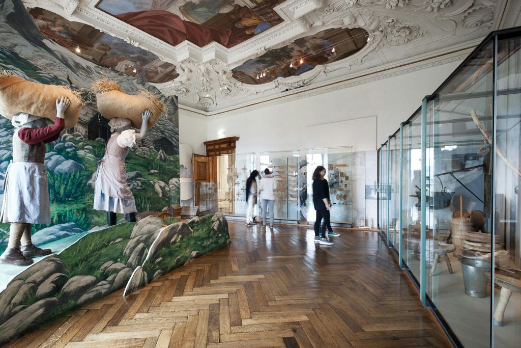 나무 바닥, 천장 벽화, 전시 케이스, 농장을 테마로 한 벽화, 전시품을 관람하는 사람들이 있는 화려한 객실입니다.