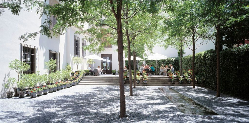 화창한 날 녹음과 나무로 둘러싸인 하얀 우산이 놓인 야외 카페 테이블에 앉아 있는 사람들.