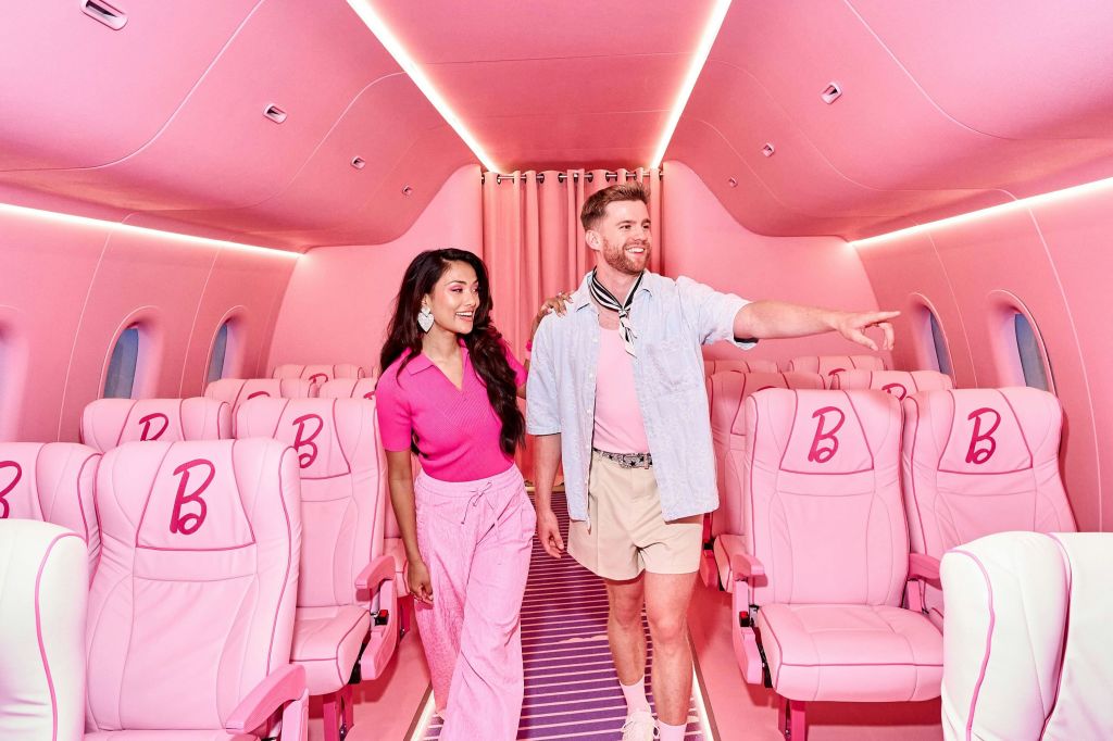 분홍색 좌석에 "B" 로고가 있는 분홍색 비행기 오두막을 걷는 두 사람이 앞을 가리키고 있습니다.