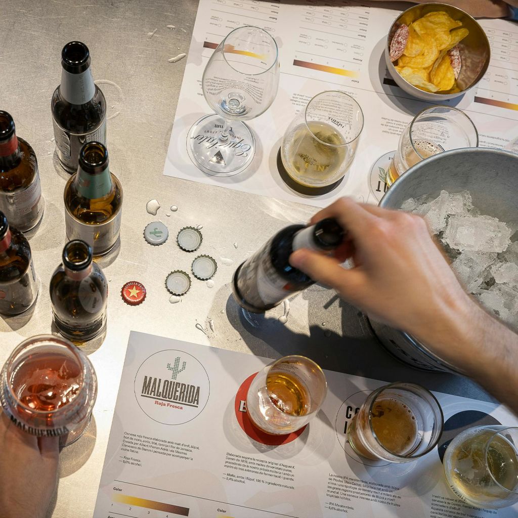 테이블 위에 맥주병 몇 개, 유리잔, 병뚜껑, 과자 한 그릇이 놓여 있습니다. 음료를 들고 있는 두 손이 보입니다.