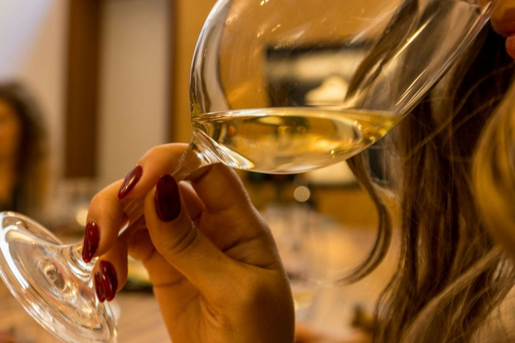 따뜻한 실내 환경에서 길고 잘 다듬어진 손톱으로 화이트 와인 한 잔을 들고 있는 사람.