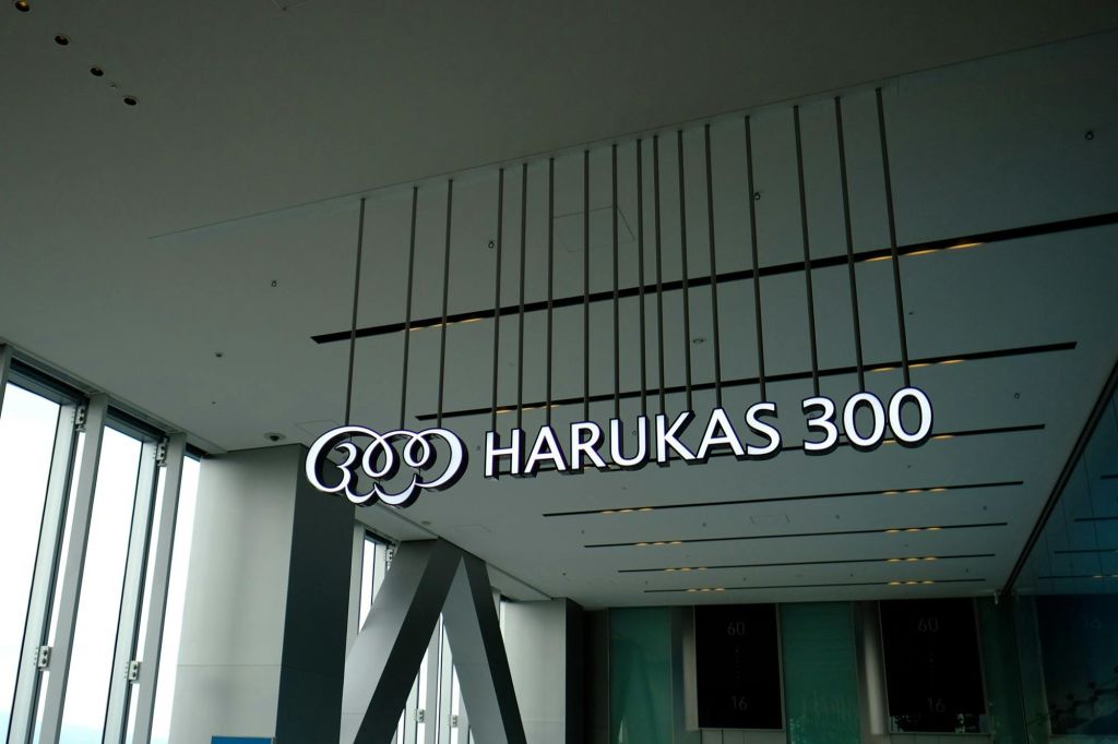 수직선이 있는 현대적인 천장에 흰색 글자로 "HARUKAS 300" 이라고 적힌 커다란 표지판이 있는 실내 공간.