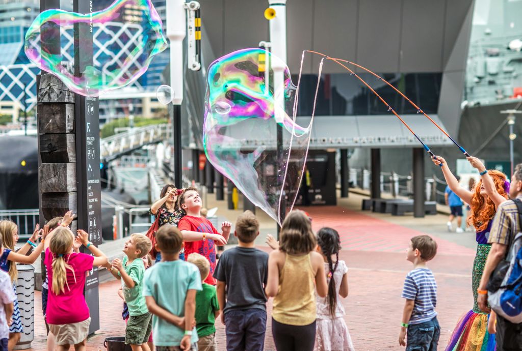 현대적인 건물과 도시적인 요소를 배경으로 야외에서 거대한 거품을 만들어내는 모습을 지켜보는 어린이들.