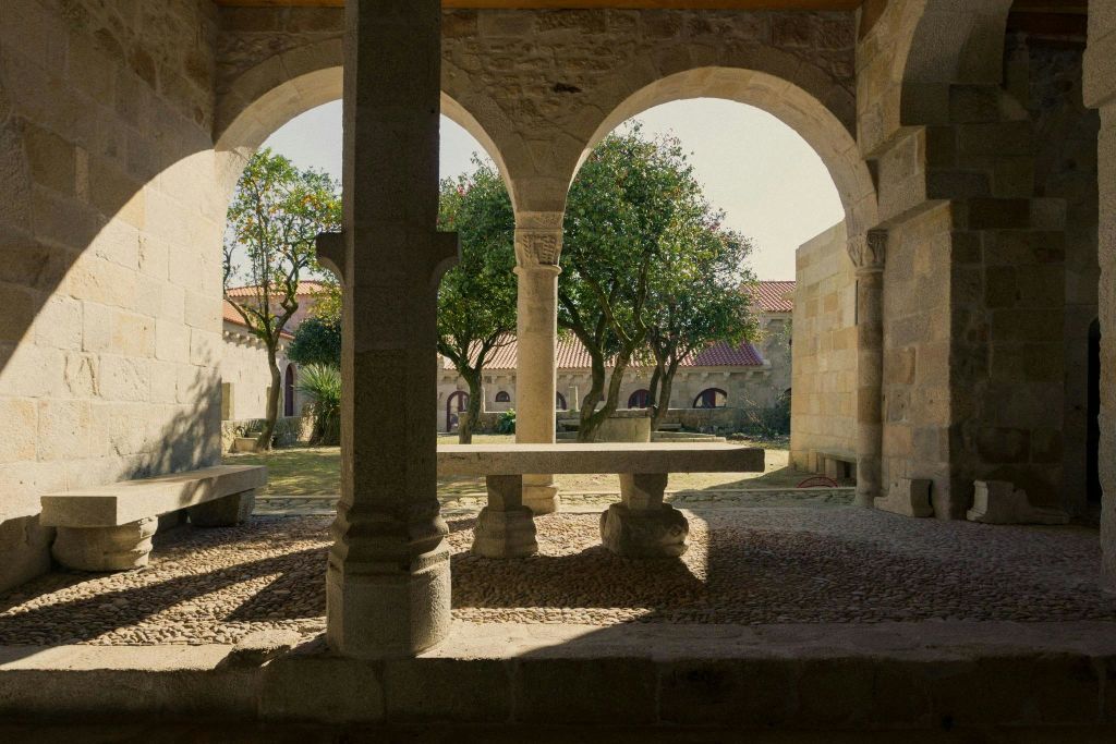 아치가 있는 돌 마당, 돌 테이블, 벤치. 나무와 붉은 지붕의 건물이 배경으로 보입니다.
