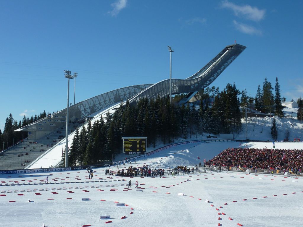 커다란 스키 점프와 눈 덮인 풍경, 나무, 스코어보드를 배경으로 맑은 하늘 아래에서 스키 경기를 관람하는 사람들.