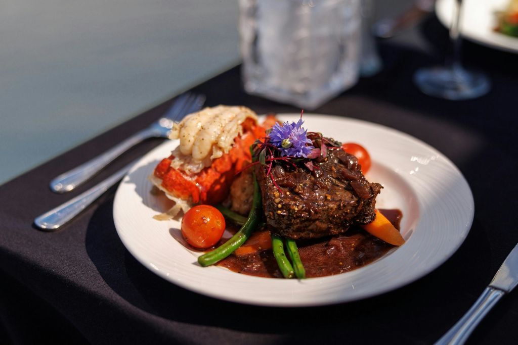 흰색 접시에 스테이크, 랍스터 꼬리, 체리 토마토, 녹두, 장식품이 담긴 고급 접시입니다.