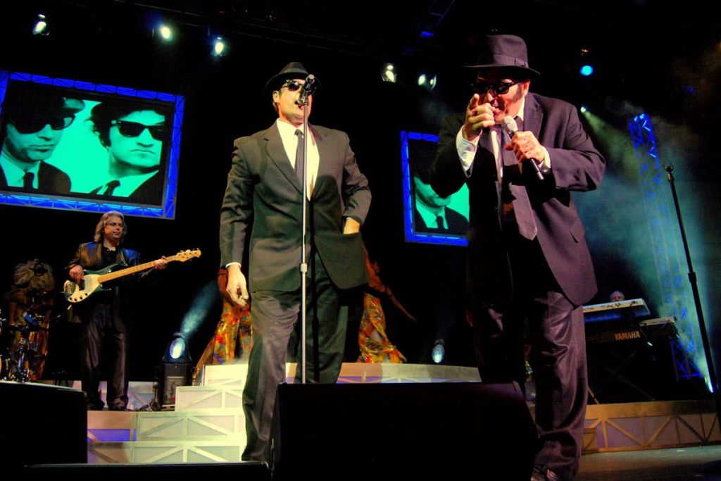 수트와 모자를 쓴 가수와 기타리스트가 함께 무대에서 공연하는 밴드. 선글라스를 쓴 두 남자의 이미지가 뒤에 표시됩니다.