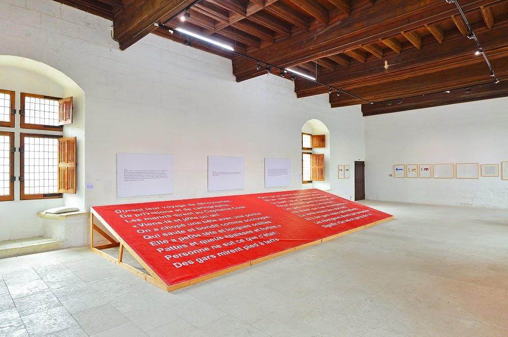 흰색 벽, 예술품, 나무 천장 기둥이 있는 넓은 방에 세워진 프랑스어로 된 흰색 텍스트가 있는 대형 빨간색 플랫폼.