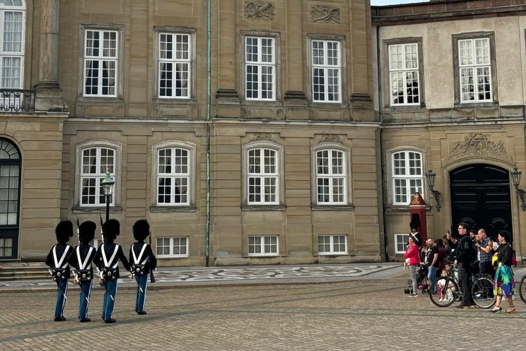 유니폼과 높은 모자를 쓴 경비원들이 유서 깊은 건물 앞에 서 있으며, 멀리서 지켜보는 사람들도 적지 않습니다.