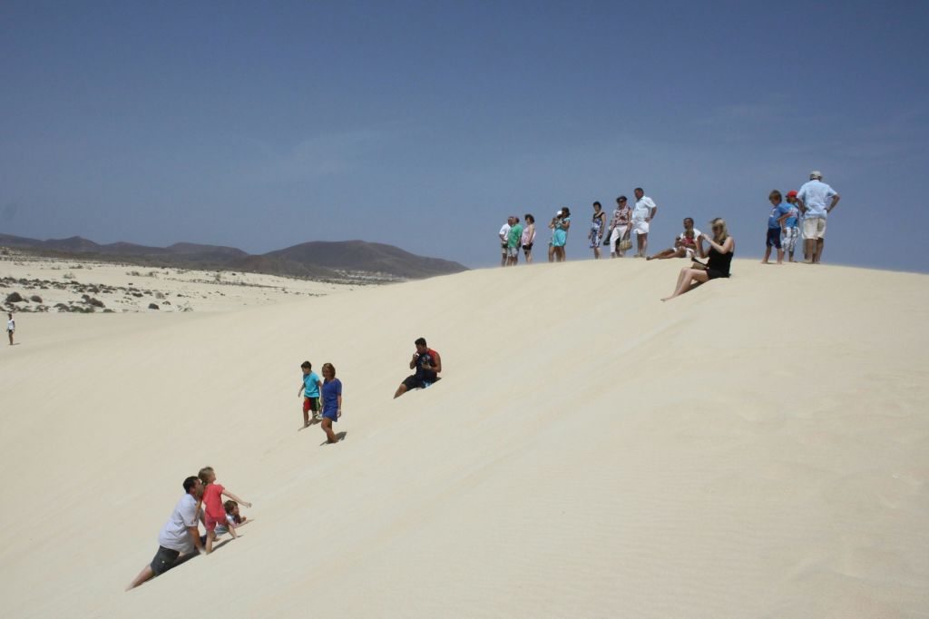 멀리 보이는 산을 배경으로 맑고 푸른 하늘 아래 커다란 모래 언덕에 사람들이 서 있습니다.