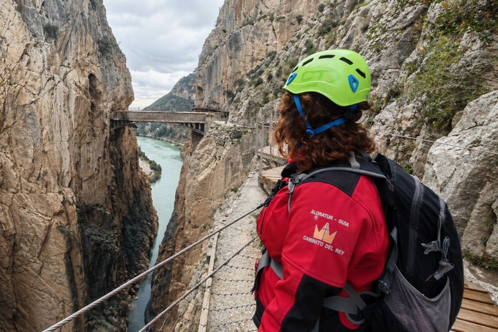 빨간 재킷과 녹색 헬멧을 입은 사람이 절벽을 따라 좁은 통로에 서서 멀리 다리가 있는 협곡을 내려다보고 있습니다.