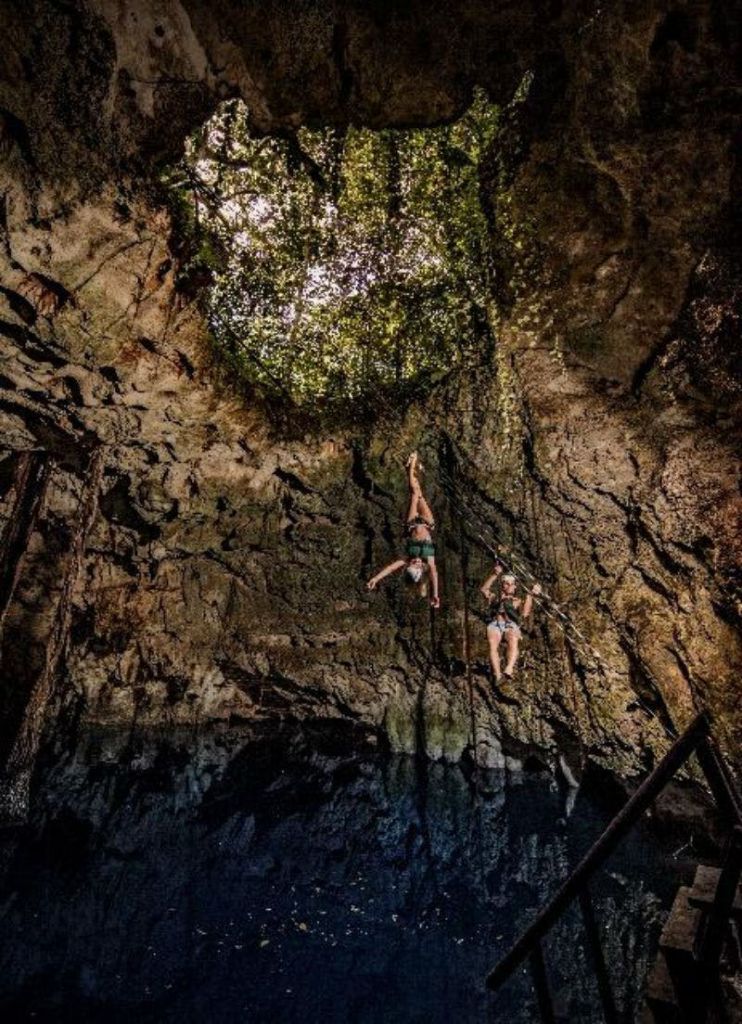 두 사람이 동굴 내부의 밧줄에 매달려 있으며, 꼭대기에 개구부가 있고, 단풍을 통과하는 햇빛 필터링으로 조명이 켜져 있습니다.