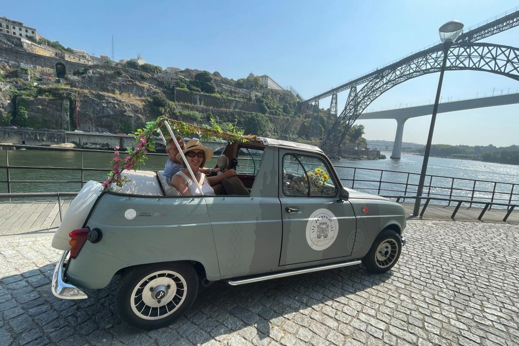 다리와 언덕에 있는 건물을 배경으로 강가에 주차된 빈티지하고 장식된 컨버터블 차량을 타고 있는 두 사람.