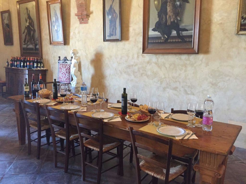벽에 나무 의자와 그림으로 둘러싸인 공간, 와인병, 물, 빵이 놓인 긴 식탁.