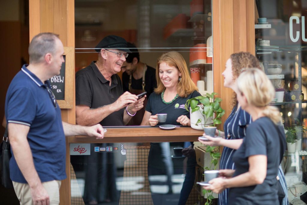 카페 밖에서 커피를 마시며 웃고 대화하는 네 명의 사람들. 카운터 뒤에서 한 남성이 여성에게 전화를 건다.
