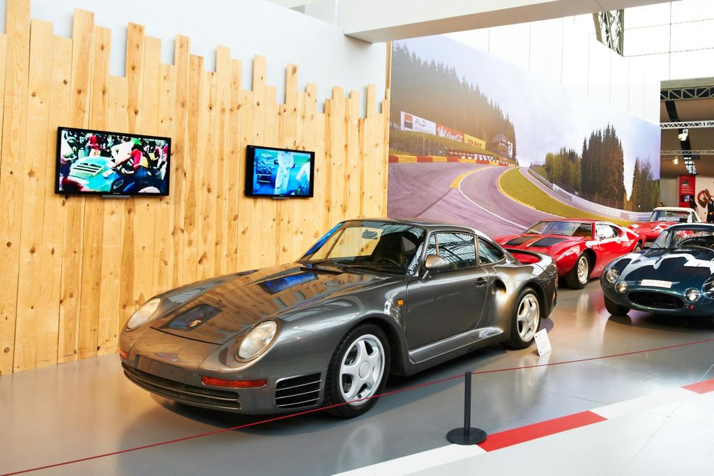 회색 스포츠카를 포함한 클래식 자동차가 전면에 전시되어 있습니다. 나무 벽과 레이싱 사진이 배경입니다.