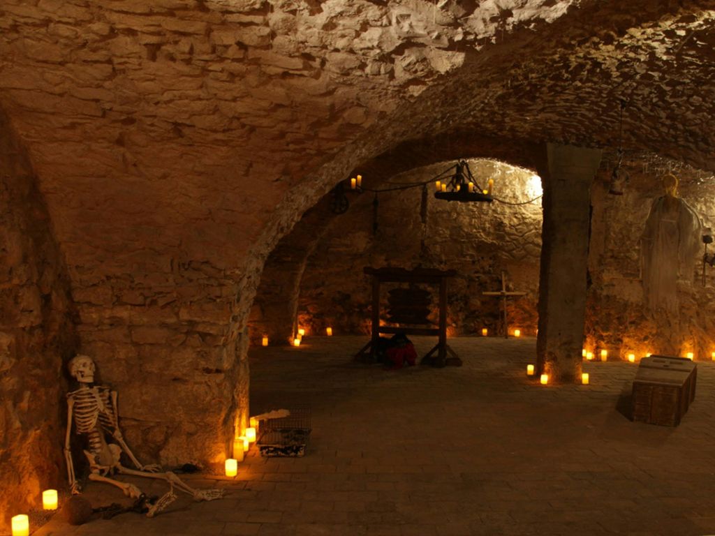 해골, 양초, 나무 테이블이 있는 조명이 희미한 석조 지하실. 배경에 십자가와 샹들리에가 보입니다.