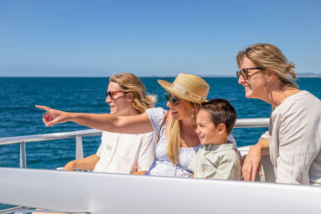 세 명의 여성과 한 명의 소년이 배를 타고 미소를 지으며 무언가를 가리키고 있습니다. 선글라스와 여름옷을 입고 맑고 푸른 바다를 배경으로 활동합니다.