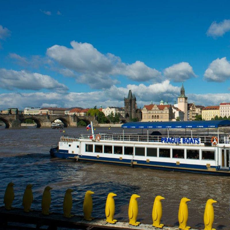 도시 건물과 돌다리를 배경으로 강에 "프라하 보트" 라고 표시된 페리, 전경의 난간에 있는 노란 펭귄 동상.