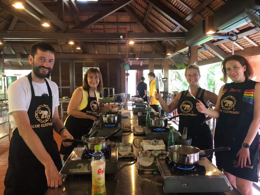 소박한 주방에서 앞치마를 두르고 조명이 걸린 높은 나무 천장 아래에서 개별 스테이션에서 요리하는 세 사람.