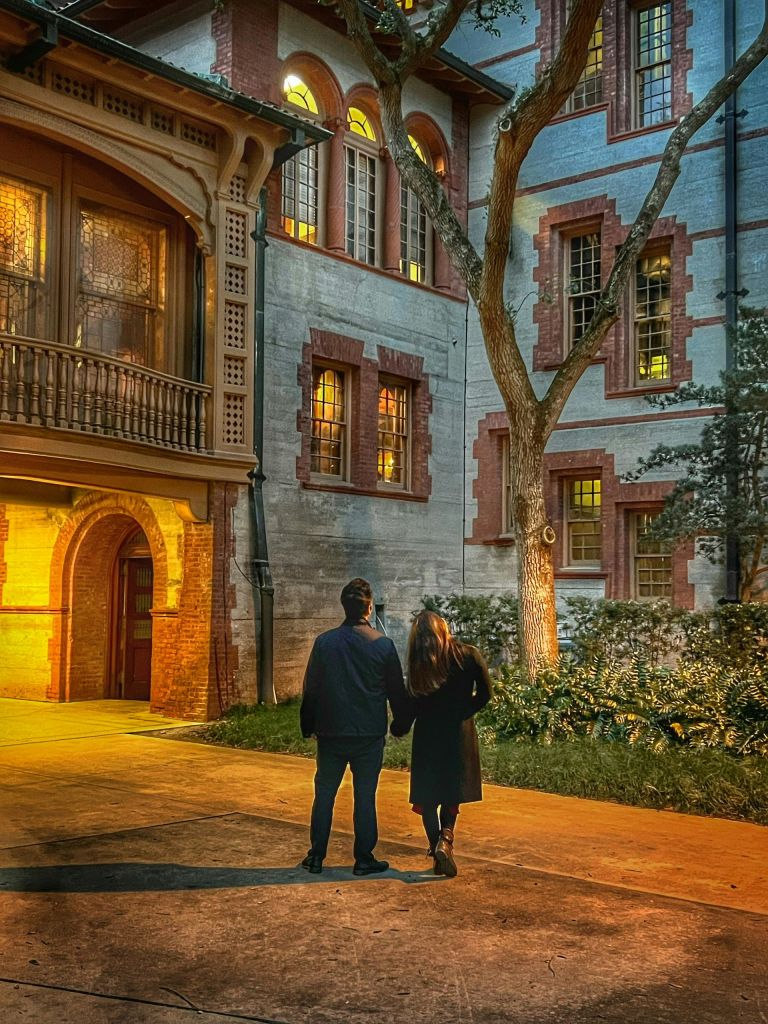 밤에 아치형 창문과 나무가 있는 유서 깊은 건물 근처에서 커플이 손을 잡고 함께 서 있습니다.