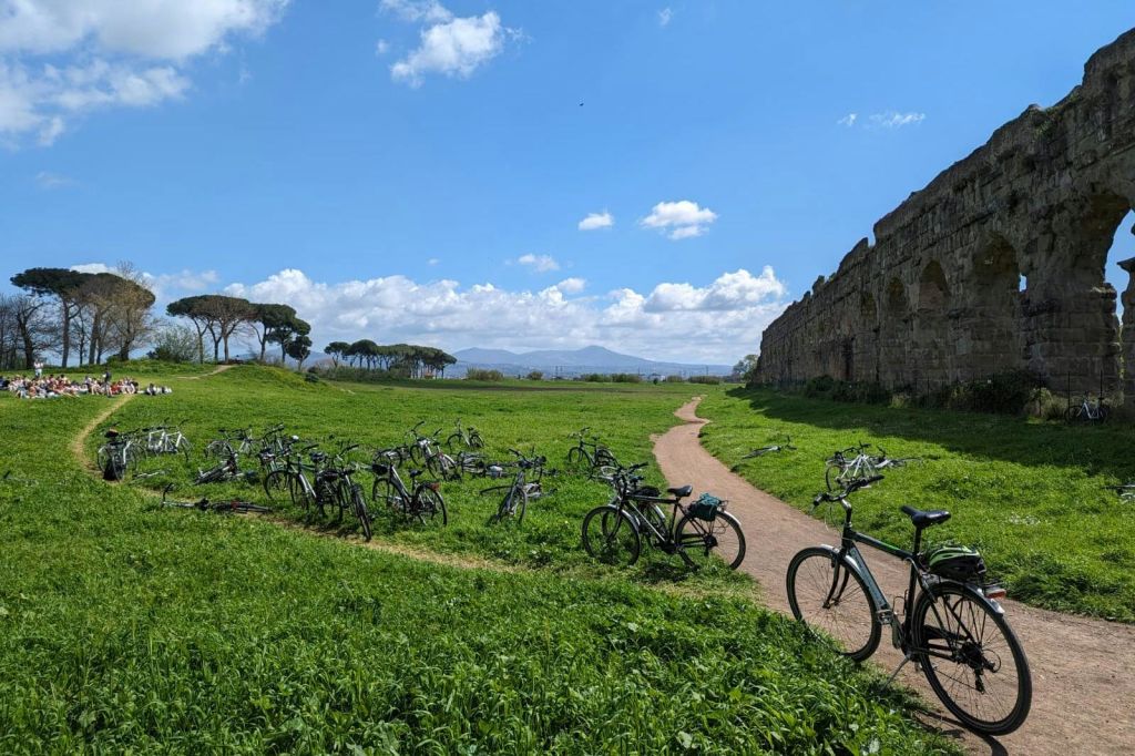 흙길, 수많은 주차된 자전거, 나무, 구름이 있는 푸른 하늘 아래 폐허가 있는 경치 좋은 잔디밭.