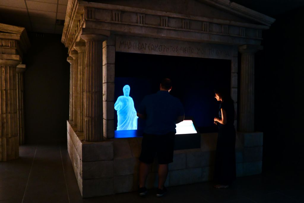 두 사람은 고대 그리스 건축의 디테일이 돋보이는 석조 건축물 내부에 있는 파란색 홀로그램 조각상을 관찰합니다.