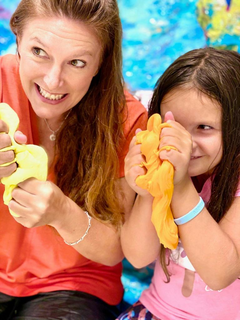 한 여성과 소녀가 다채롭고 흐릿한 배경의 노란색과 주황색 점액으로 장난을 치며 웃고 있습니다.