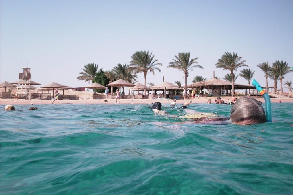 초가 우산과 야자수가 있는 해변, 맑고 푸른 물이 흐르는 해변 근처에서 수영하고 휴식을 취하는 사람들.